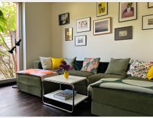 Gemütliches Wohnzimmer mit grünem Ecksofa, bunten Kissen und Decken sowie einem weißen Couchtisch mit Vase; an der Wand hängt eine Galerie aus gerahmten Bildern. Links sorgt eine große Fensterfront mit Blick ins Grün und eine Stehlampe für viel Tageslicht.