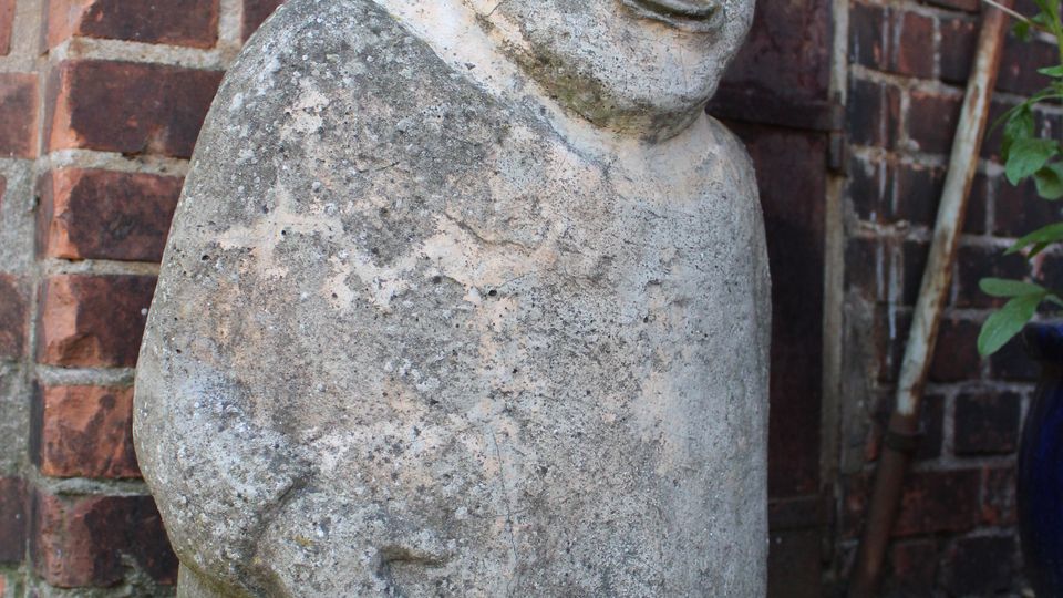 Verwitterte Steinfigur in Zwergform mit spitzer Mütze vor einer roten Backsteinmauer. Die Figur steht auf einem Betonboden, daneben sind einige Pflanzen und ein schmaler Holzpfahl zu sehen.