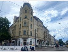 Eckgebäude mit heller Fassade im Gründerzeitstil, mit Erkern und einem spitz zulaufenden Dach. Im Vordergrund Straßenbahnschienen, Stromleitungen sowie ein Baustellenbereich mit Absperrungen und Baugeräten.