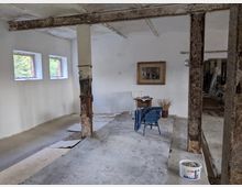 Ein unfertiger Innenraum mit weißen Wänden, grobem Betonboden und sichtbaren, rostigen Metallstützen. An der Wand hängt ein gerahmtes Bild, und im Raum stehen vereinzelt Möbel, darunter ein Tisch, zwei Stühle und ein kleiner Eimer auf dem Boden. Links sind drei rechteckige Fenster, die Tageslicht hereinlassen.