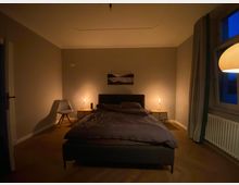 Ein Schlafzimmer mit gedämpfter Beleuchtung. Ein Doppelbett steht mittig vor einer beigen Wand, flankiert von zwei kleinen Nachttischen mit leuchtenden Tischlampen. Rechts befindet sich ein Fenster mit Vorhängen, links ein grauer Stuhl neben einer Tür.