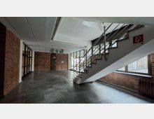Innenbereich eines Treppenhauses in einem Gebäude mit Backsteinwänden, dunkelgrauem Steinboden und weißer Decke. Eine Treppe mit Metallgeländer führt nach oben, daneben große Fensterfronten und Glastüren entlang des Flurs.