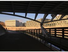 Blick unter einer Brücke auf moderne Bürogebäude mit rechteckiger Fensterfassade entlang eines Flussufers. Im Vordergrund verläuft ein Fußweg mit Geländer, während die Gebäude im Hintergrund in der Abendsonne beleuchtet sind.