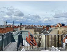 Blick von einer Dachterrasse über die Dächer einer Stadt mit einer Mischung aus älteren Gebäuden mit roten Ziegeldächern und modernen Hochhäusern im Hintergrund. Der Himmel ist bewölkt, und im Vordergrund sind ein Geländer und Teile der Terrasse erkennbar.