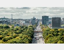Städtische Panoramaansicht mit Blick auf eine breite, von Bäumen gesäumte Straße, die in die Ferne führt, flankiert von modernen Gebäuden und Hochhäusern. Im Hintergrund sind der Berliner Funkturm und andere städtische Strukturen zu sehen.