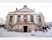 Historisches Gebäude mit klassizistischer Fassade, seitlicher Treppe und Sandsteinverzierungen, darüber ein Dreiecksgiebel mit Inschrift. Im Vordergrund ein Eingangsbereich mit geöffneten Doppeltüren und moderner Beschilderung.