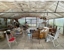 Ein rustikales, überdachtes Gewächshaus mit transparenten Wänden und einer Stoffplane als Dach. Im Raum stehen verschiedene Sitzmöbel, wie Stühle und Sofas, die ungeordnet um Tische gruppiert sind; im Hintergrund sind getrocknete Pflanzen und Dekorationen aufgehängt.