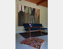 Ein Wohnzimmer mit einem Mid-Century-Sofa in Blau, einem passenden Holz-Couchtisch und einem dunklen Teppich. Im Hintergrund hängt ein großformatiges Gemälde mit gezeichneten Männeranzügen, und im Vordergrund liegt ein bunt gemustertes Bodenkissen.