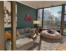 Wohnzimmer mit großen Fenstern und Jalousien, das viel Tageslicht hereinlässt. Ein graues Sofa mit bunten Kissen steht vor einer dunkelgrünen Wand, an der eine Neonlampe mit der Aufschrift „love“ und ein großes Schwarz-Weiß-Foto hängen. Im Raum befinden sich ein flauschiger Sitzsack, ein Teppich und ein Couchtisch mit verschiedenen Gegenständen.