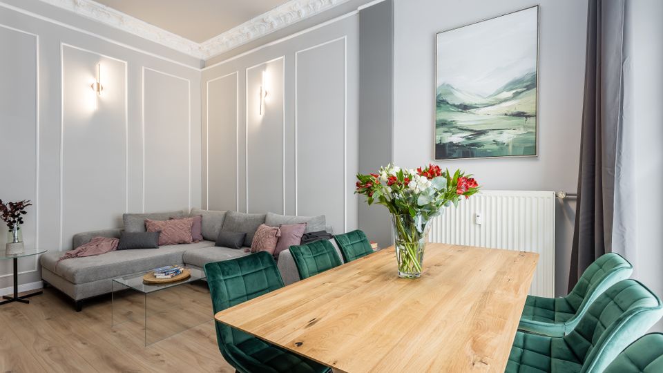 Heller, modern eingerichteter Wohn- und Essbereich mit grauen Wänden samt Stuckleisten und Wandpaneelen, einem Ecksofa und einem Holzesstisch mit grünen Polsterstühlen. An der Wand hängen ein Landschaftsbild und darunter ein Heizkörper, daneben ein großes Fenster mit dunklen Vorhängen.