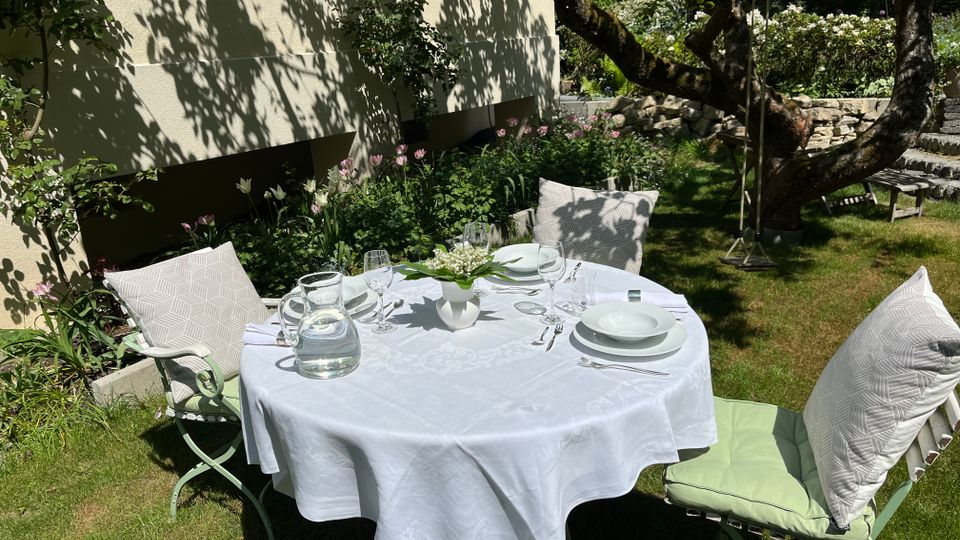 Runder Gartentisch mit weißer Tischdecke und gedecktem Geschirr steht auf einer Rasenfläche neben der Hausfassade mit Fenstern und einem Blumenbeet. Im Hintergrund sind ein großer Baum, eine niedrige Natursteinmauer und weitere Gartenpflanzen zu sehen.