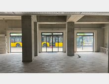 Rohbau eines Erdgeschosses mit unverputzten Betonwänden, großen Fenstern und einem gläsernen Eingang mit automatischen Schiebetüren. Draußen ist ein gelber Linienbus sichtbar, der an einer Haltestelle vorbeifährt.