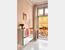 Wohnraum mit hellen Wänden, einer verzierten Stuckdecke und einem offenen Fenster, das auf einen Balkon mit roten Gartenstühlen und Pflanzen in Töpfen führt. Ein schwebendes, verspiegeltes Sideboard steht an der Wand, darüber hängt ein abstraktes Gemälde.