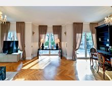 Helles Wohnzimmer mit Holzboden im Fischgrätenmuster, großen Fenstern mit beigen Vorhängen und einem Glasgang zum Außenbereich. Links ein Fernseher auf einem weißen Schrank, rechts ein Bücherregal und ein eleganter Schreibtisch mit gepolsterten Stühlen.