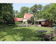 Großer Garten mit Rasenfläche, darauf mehrere Holztische und Stühle, eine Holzbank sowie ein runder Tisch im Vordergrund. Im Hintergrund steht ein gelbes Haus mit roten Ziegeldach und grünen Fensterläden, daneben ein Holzschuppen und ein Trampolin, umgeben von vielen Bäumen.