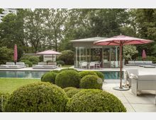 Moderner Garten mit Pool, umgeben von gepflegten Buchsbaumsträuchern und einer Terrasse mit Sonnenliegen und Sonnenschirmen. Im Hintergrund ein verglaster Pavillon mit Sitzgelegenheiten und dichter Baumbewuchs.
