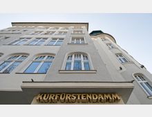 Fassade eines mehrstöckigen Gebäudes mit hellen Putzwänden und großen Fenstern im klassischen Stil. Unten ist der Schriftzug „KURFÜRSTENDAMM“ in goldenen Buchstaben angebracht.