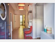 Moderner Flur mit Holzfußboden, weißen Wänden und roten Deckenlampen. Links befindet sich eine offene Tür mit einer runden Glasscheibe, geradeaus führt ein heller Gang zu einem Raum mit Pflanzen, rechts stehen ein roter Stuhl und dekorative Kerzen auf einem Sideboard.