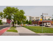 Straßenszene mit Gehweg, Fahrradweg und einer Fahrbahn, rechts ein Backsteingebäude mit einem Netto-Supermarkt und geparkten Autos, dahinter mehrstöckige Wohngebäude. Links steht ein großer Baum neben einer roten Schule mit der Aufschrift „Oberschule“.