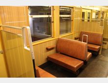 Innenansicht eines alten Zugabteils mit Holzwänden und braunen Kunstledersitzen. Die Fenster sind mit Metallrahmen versehen, und seitlich befinden sich Haltegriffe aus Metall sowie kleine Ablageflächen.