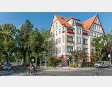 Straßenecke mit einem mehrstöckigen Wohnhaus im Stil eines Altbaus, gekennzeichnet durch helle Fassade, rote Dachziegel und Balkone mit Blumenkästen. Die Umgebung zeigt eine Baumallee, parkende Autos und einen Radfahrer auf der Straße, Straßenlaternen und Verkehrsschilder ergänzen die Szene.