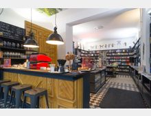 Innenaufnahme eines gemütlichen Ladens, der eine Kombination aus Café und Buchladen darstellt. Im Vordergrund eine Theke mit Holzverkleidung, Barhockern, einer roten Kaffeemaschine sowie Gebäck; im Hintergrund Regale voller Bücher und Geschenkartikel, umgeben von weiß gestrichenen Wänden und dekorativen Deckenleuchten.