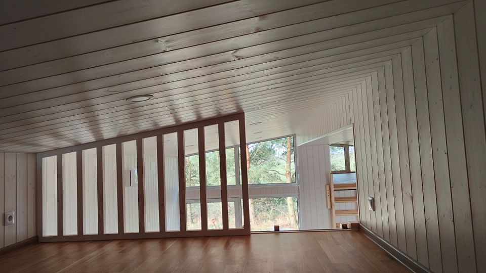 Dachgeschossraum mit schräger Holzdecke und holzverkleideten Wänden, Holzfußboden sowie einer niedrigen Trennwand mit senkrechten Streben. Große Fensterfront mit Blick auf Bäume; rechts ein kleiner Durchgang zu einem tieferen Bereich mit Treppe.