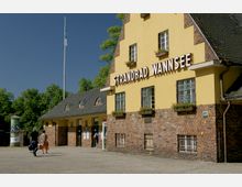 Gelb gestrichenes Gebäude mit rotem Ziegelsockel und einem Satteldach, beschriftet mit „Strandbad Wannsee“. Vor dem Eingang gehen Personen, im Hintergrund sind Bäume und ein Fahnenmast sichtbar.