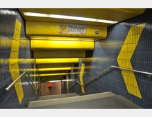 Treppenabgang einer U-Bahn-Station mit blauen Kacheln an den Wänden, die durch gelbe Streifenmuster akzentuiert sind. Die Decke besteht aus gelben Paneelen, und ein Schild mit der Aufschrift „Nichtraucher Zone“ ist sichtbar.