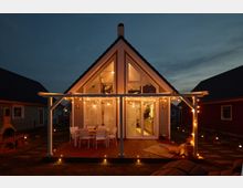 Moderner, beleuchteter Ferienhaus-Bungalow im A-Frame-Stil mit großen Glasfronten und einer Holzterrasse, dekoriert mit Lichterketten und Gartenmöbeln, in einer ruhigen Nachbarschaft bei Abenddämmerung.