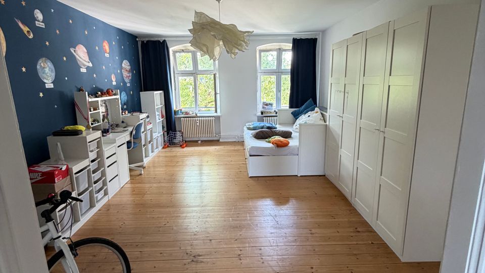 Heller Wohnraum mit breitem Holzdielenboden, zwei großen Fenstern mit dunklen Vorhängen und einer Deckenlampe. Links eine dunkelblaue Wand mit Weltraum-Tapete und Regalen, rechts eine Reihe weißer Kleiderschränke sowie ein weißes Tagesbett vor den Fenstern.