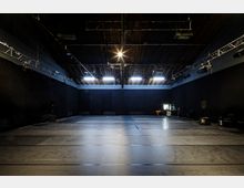 Ein leerer, großzügiger Raum mit dunklen Wänden und einem Holzdach, ausgestattet mit Beleuchtungs- und Bühnentechnik, einschließlich Scheinwerfern und Traversen. Der Boden ist eben und dunkel, seitlich stehen technische Geräte und Möbel.