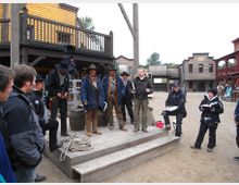 Szene in einer Westernstadt mit Holzbauten im Hintergrund. Eine Gruppe von Personen, darunter mehrere in Westernkleidung, steht vor einer erhobenen hölzernen Plattform neben einem Galgen, umgeben von Kameratechnik und Crew-Mitgliedern.