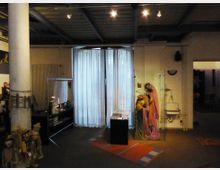 Ein Raum mit dimmer Beleuchtung, im Vordergrund ein Podest mit Broschüren, rechts eine Glasvitrine mit einer Figurengruppe in bunten Gewändern. Im Hintergrund bedecken weiße Vorhänge große Fenster, während links Puppen und Regale zu sehen sind.