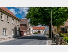 Eine ruhige Dorfstraße mit zweigeschossigen Wohnhäusern, teils aus Putz und teils aus Backstein. Im Hintergrund steht ein weißes Gebäude mit rotem Ziegeldach und einem großen Torbogen, umgeben von Bäumen und einem Gehsteig mit Laternen.