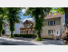 Straßenszene mit mehreren Wohnhäusern in einer Reihe, darunter ein historisches Backsteingebäude mit Giebeldachfenstern und ein schlichtes verputztes Haus mit rechteckigen Fenstern. Vor den Häusern stehen große, schattenspendende Bäume und Büsche, daneben verläuft ein Gehweg.