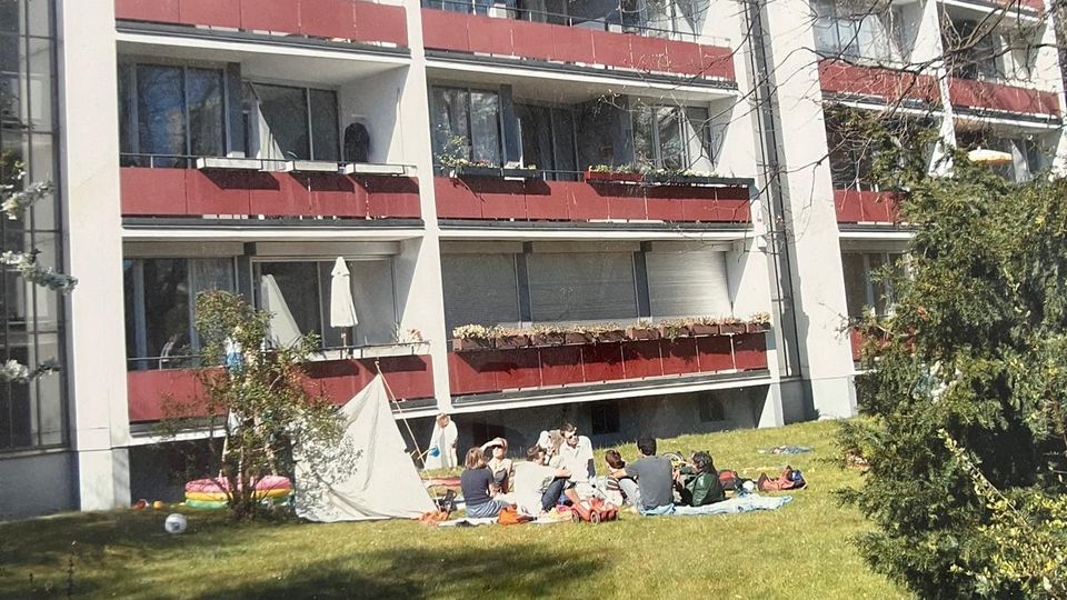 Mehrstöckiges Wohnhaus mit weißer Fassade und durchgehenden Balkonen mit roten Brüstungen. Davor liegt eine große Rasenfläche mit Sträuchern und Bäumen am Rand unter blauem Himmel.
