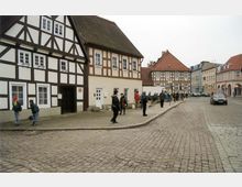 Eine gepflasterte Straße in einer Altstadt mit Fachwerkhäusern im Vordergrund. Menschen stehen und gehen entlang des Gehwegs, und im Hintergrund sind weitere historische Gebäude mit Satteldächern sichtbar.
