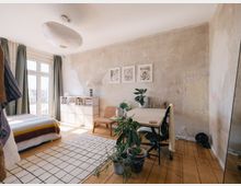 Helles Schlafzimmer mit einem Schreibtisch und einem Bett. Der Raum hat eine unverputzte Wand, Holzboden, große Fenster mit Vorhängen, Zimmerpflanzen und minimalistische Möbel.