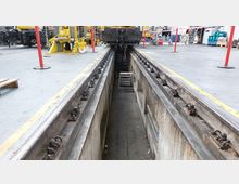 Eine industrielle Werkhalle mit einem Wartungsgleis, das in einen tiefen Betonschacht eingebettet ist. Im Hintergrund sind Werkzeuge, Ausrüstung und ein Schienenfahrzeug sichtbar.