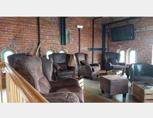 Gemütlicher Aufenthaltsraum mit braunen Ledersesseln und passenden Fußhockern auf Holzboden. Die Wände bestehen aus rotem Sichtmauerwerk mit kleinen, rundbogigen Fenstern, und ein großer Fernseher hängt an der Wand.