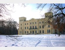 Schlossartiges Gebäude mit cremefarbener Fassade und zwei kleinen Türmen, umgeben von einem schneebedeckten Park mit kahlen Bäumen. Im Vordergrund sind Fußspuren im Schnee zu sehen.