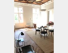 Helles Wohnzimmer mit Fischgrätenparkett, weißen Wänden und großen, bodentiefen Fenstern. Eine Holzdecke mit dekorativen Elementen und Gemälden, ein runder Esstisch mit Stühlen, ein Teppich sowie moderne und klassische Möbel prägen den Raum.