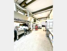 Große, moderne Großküche mit Edelstahl-Arbeitsflächen, Kochgeräten und einem Belüftungssystem an der Decke. Im Hintergrund sind Regale mit Küchenutensilien und ein Bogenfenster sichtbar.