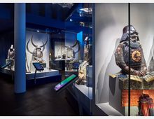 Ausstellungsraum in einem Museum, der traditionelle japanische Samurai-Rüstungen in Glasvitrinen zeigt. Die Umgebung ist dunkel mit blauem Licht, und interaktive Bildschirme ergänzen die Präsentation.