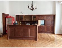 Holzvertäfelter Tresen in einem klassischen Innenraum mit hellem Parkettboden, dahinter Holzschränke mit Glastüren. Links steht ein roter Kühlschrank, an der Decke hängt ein Kronleuchter.