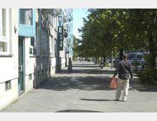 Straßenansicht mit einem Bürgersteig entlang eines Wohngebäudes mit geöffneten Fenstern und Satellitenschüsseln; rechts sind Bäume und parkende Autos zu sehen, im Vordergrund läuft ein Mann mit einer Plastiktüte.