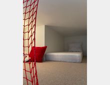 Ein niedriger Raum mit Teppichboden, einem weißen Matratzenlager samt weißem und rotem Kissen sowie einem seitlich gespannten roten Netz. Der Raum wirkt schlicht und minimalistisch eingerichtet.