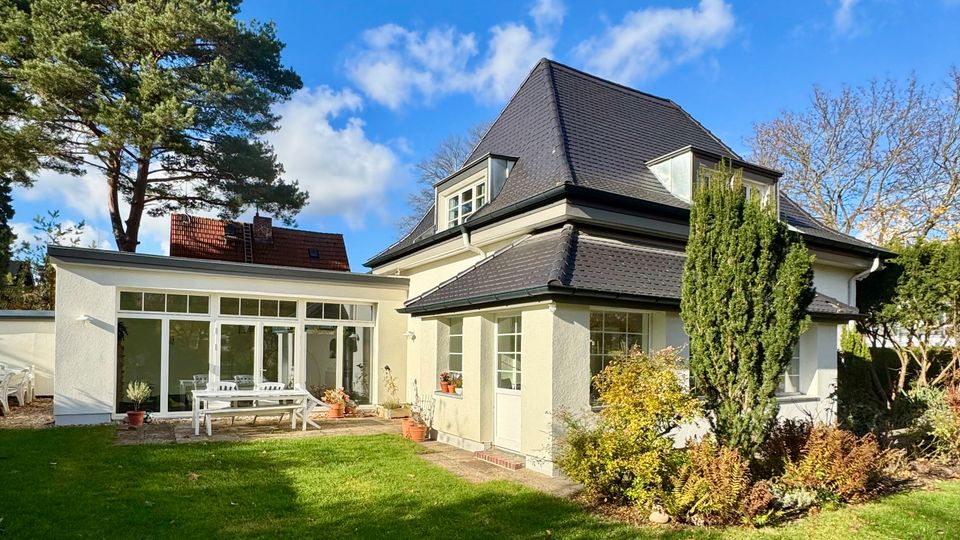 Einfamilienhaus mit dunkel gedecktem Walmdach und weißer Fassade, angrenzend an einen flachen Anbau mit großer Glasfront und Terrasse. Im Vordergrund liegt ein gepflegter Rasen mit Sträuchern und hohen Bäumen unter blauem Himmel.