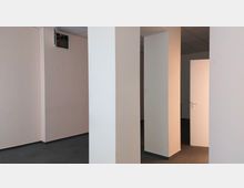 Ein leerer Innenraum mit weißen Wänden, mehreren rechteckigen Säulen und einer teilweise geöffneten weißen Tür. Der Boden ist mit grauem Teppich ausgelegt, und die Decke hat ein abgehängtes Rasterdesign.